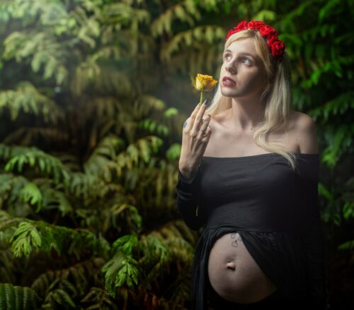 Elegant pregnant woman in a Maternity Boudoir setting, adorned with a red floral headpiece, contemplating a yellow rose amidst lush ferns.